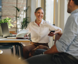 Middle-aged businesswoman holding smartphone and talking to coworker in office setting
