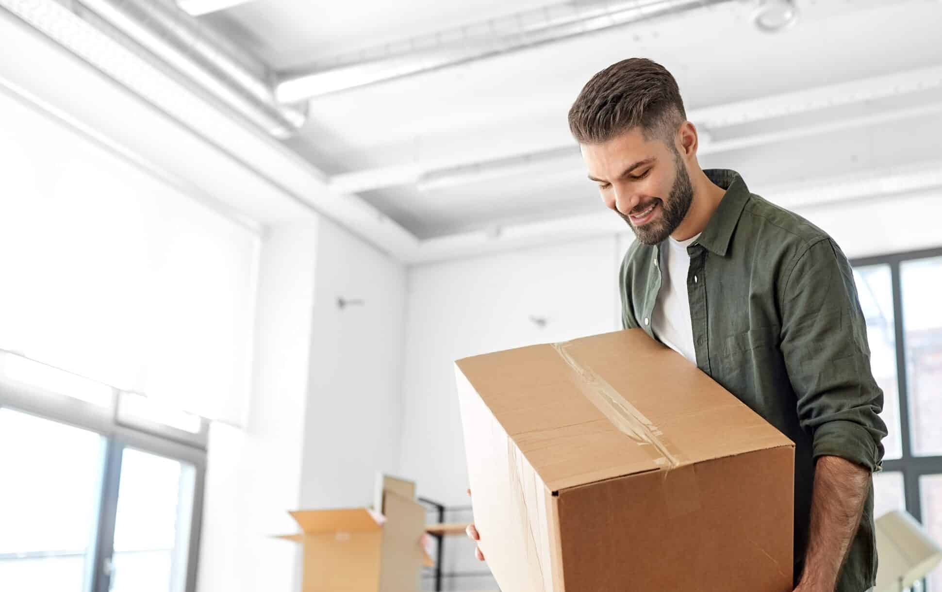 Happy smiling young man carrying moving box into new home