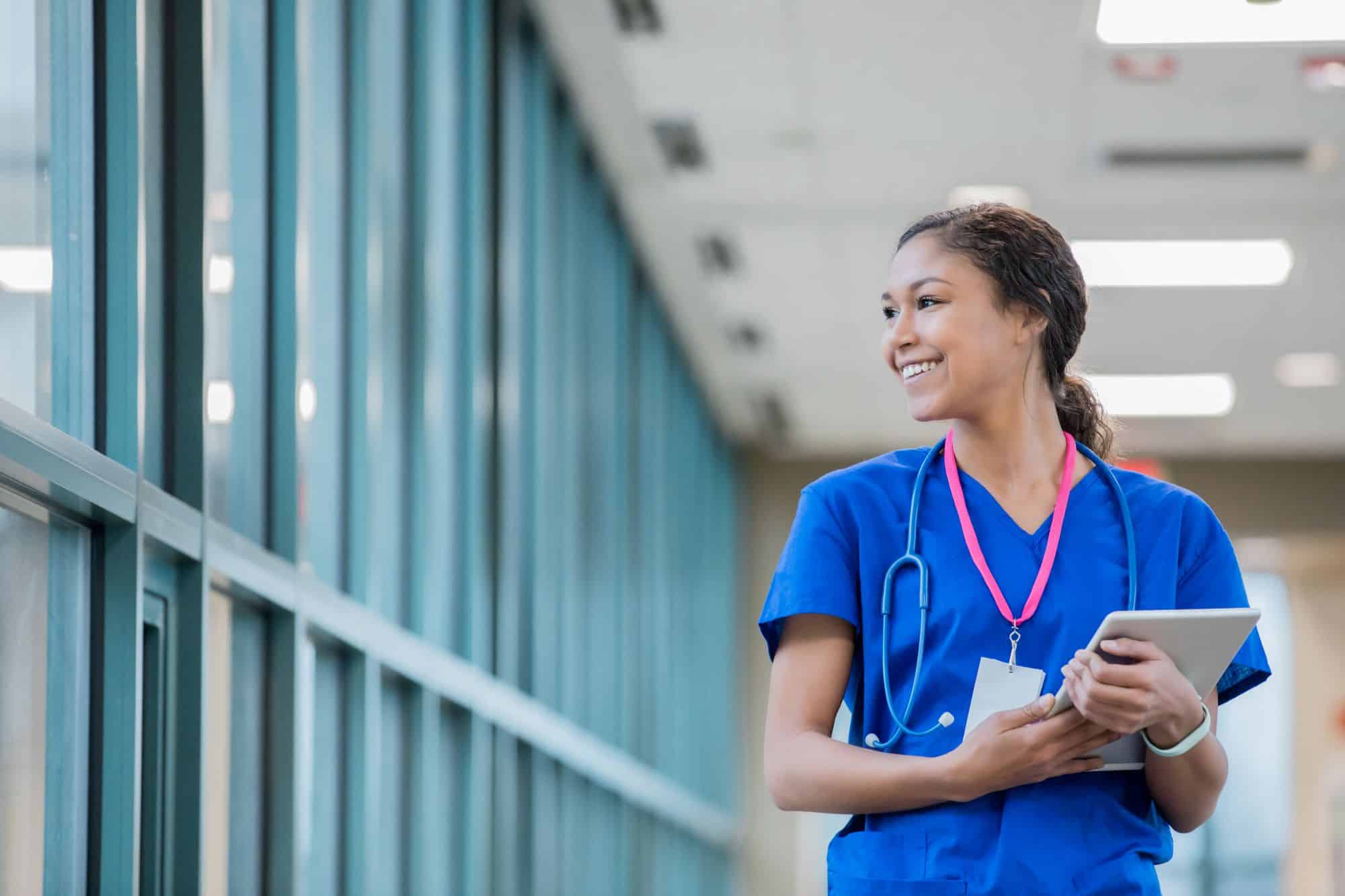Young nurse wearing stethoscope, carrying tablet, walking through hospital hallway