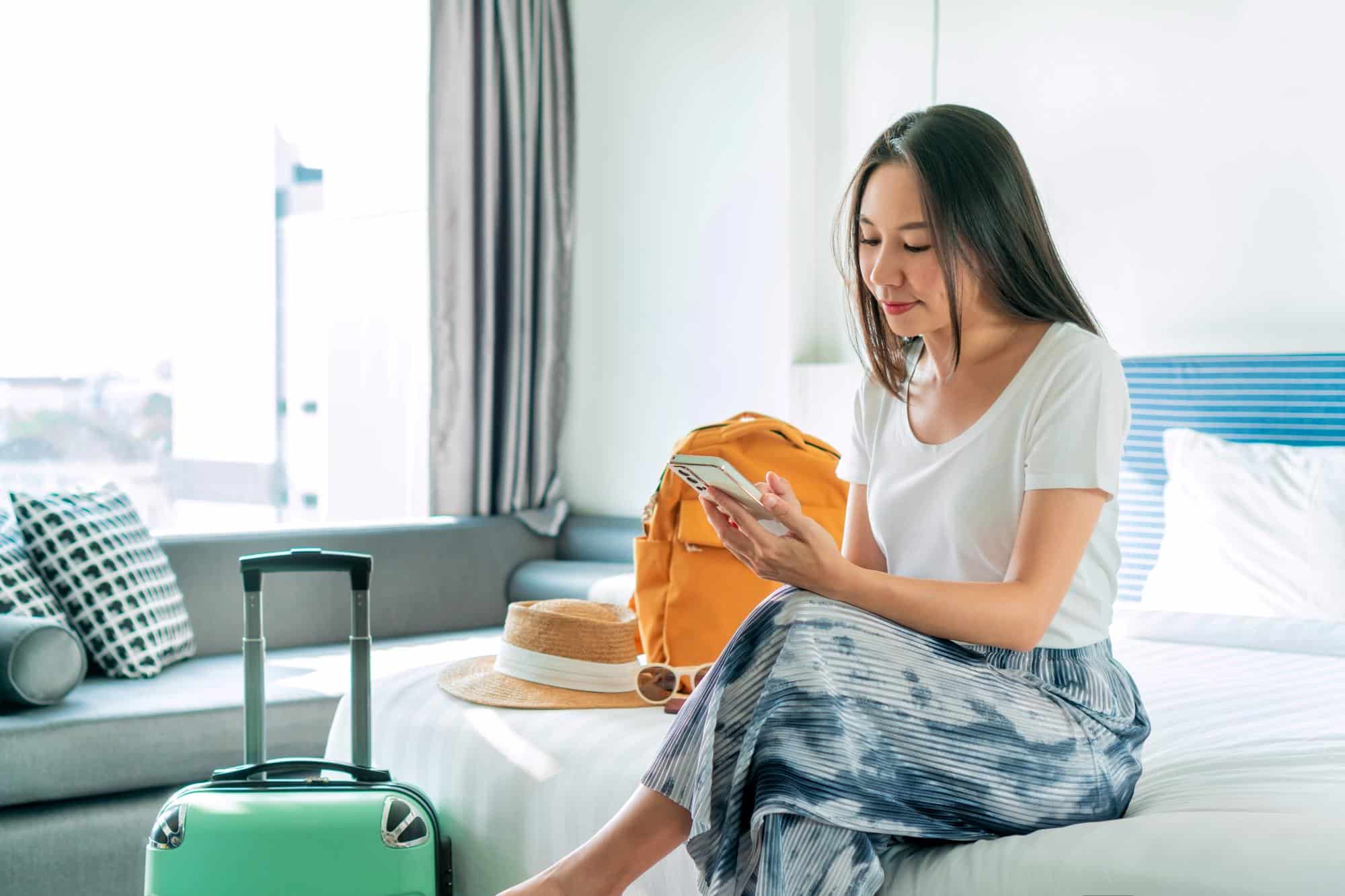 Happy young Asian traveler woman using mobile phone while relaxing on bed in hotel room next to suitcase and backpack