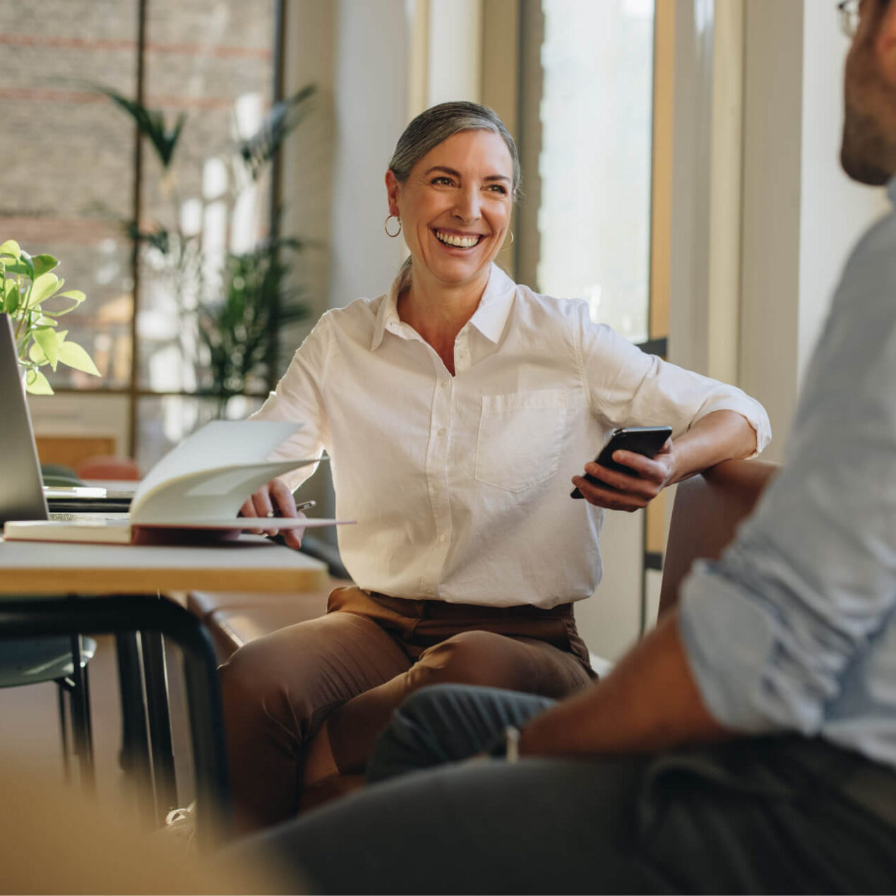 Middle-aged businesswoman holding smartphone and talking to coworker in office setting